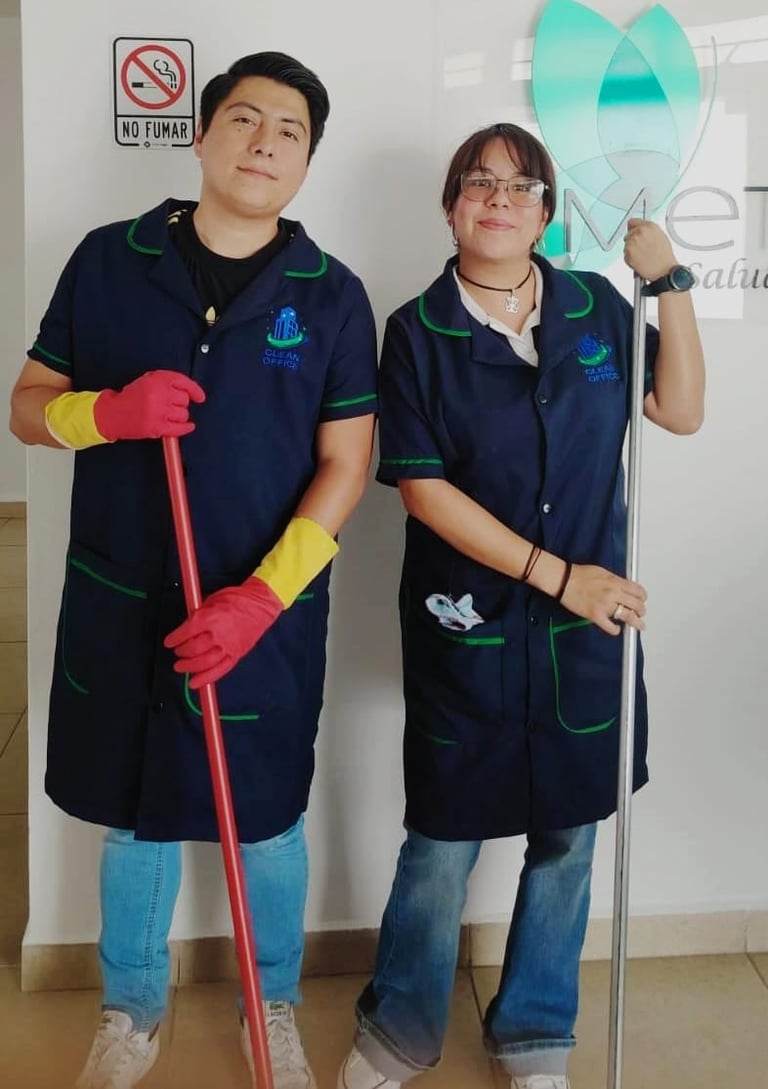 Two cleaning service workers in navy uniforms with green trim wearing yellow and red gloves, holding cleaning tools in an office space